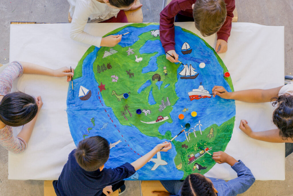 An aerial view of a multi ethnic group of children painting the globe and it's animals, fish, and vehicles. One girl paints a steam boat, while a boy colours in the ocean.