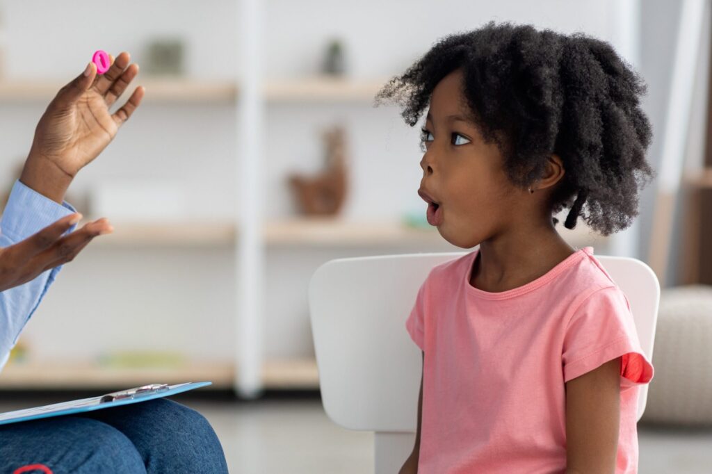 Attractive young african american woman speech therapist working with little black girl, sitting at table with colorful letters, learning pronunciation, clinic interior. Speech disorders in children
